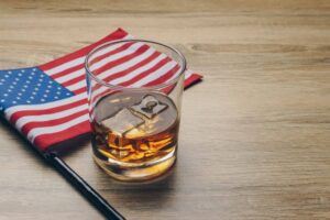 A glass of American whiskey with ice next to an American flag, representing what is American whiskey.