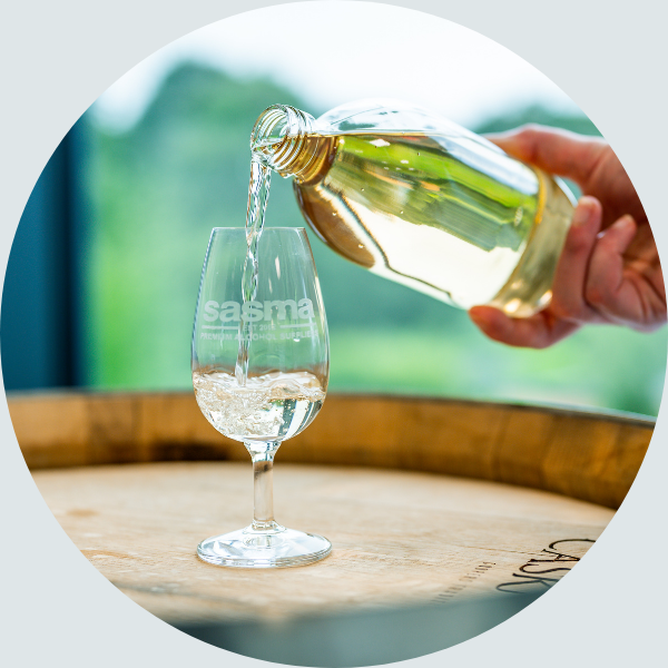 A hand pours amber liquid from a glass bottle into a Sasma-branded snifter glass