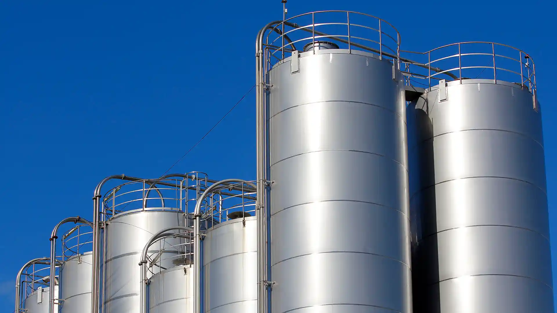 Silver industrial silos stand tall against a clear blue sky. The metal structures have railings at the top, conveying a sense of modern industry and strength.