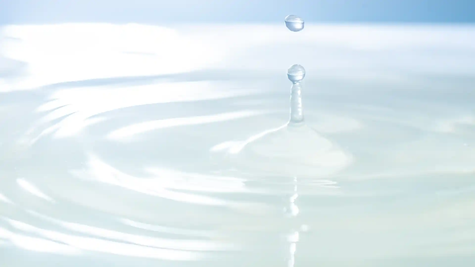 A clear droplet of water is captured mid-air above a smooth, rippling surface. The soft blue and white tones create a calming and serene atmosphere.
