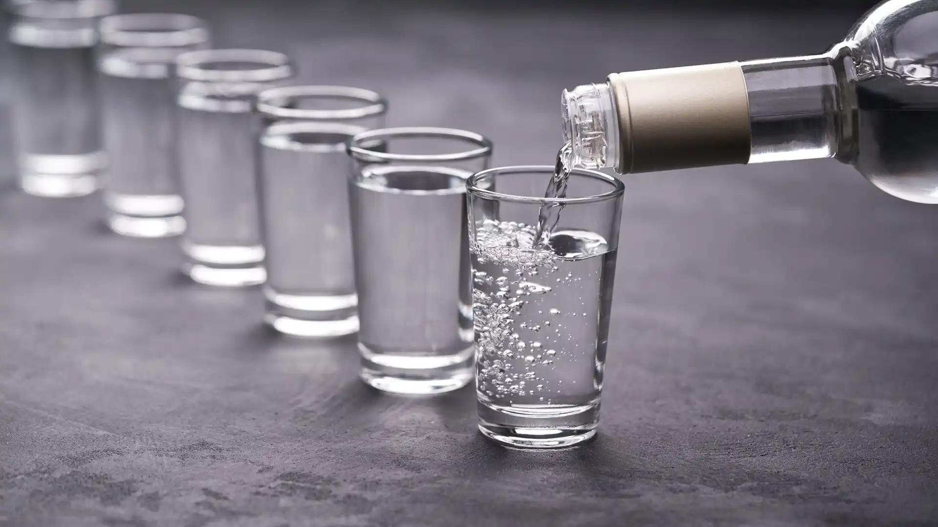 Pouring clear liquid into a row of six glasses on a dark, textured surface. The image conveys uniformity and precision with a calm, neutral tone.