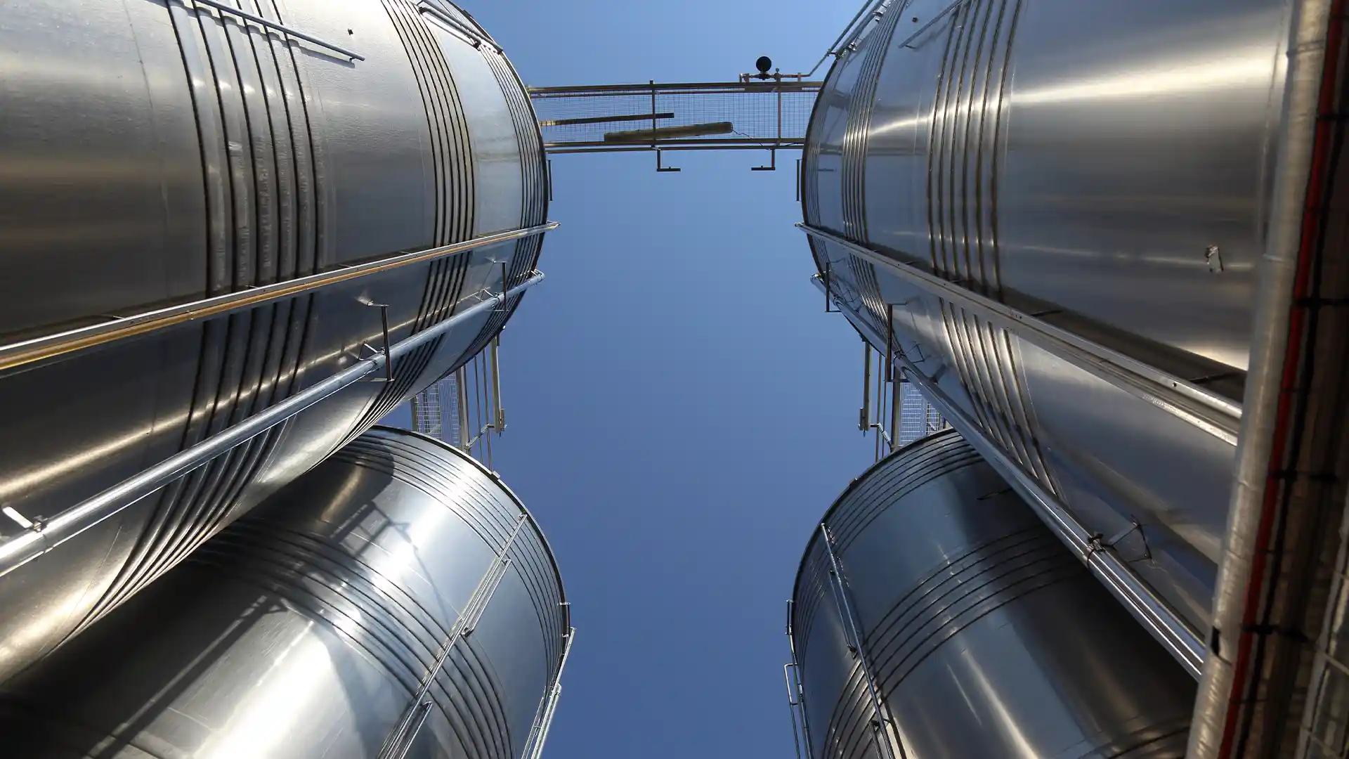 Looking up between tall, shiny metal silos under a clear blue sky, creating a sense of industrial scale and open space.