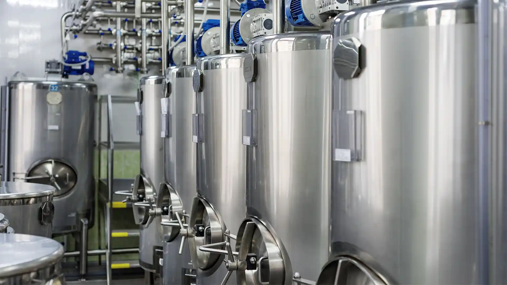 A row of stainless steel fermentation tanks with connected piping and blue motorized valves, creating a sleek, industrial atmosphere.