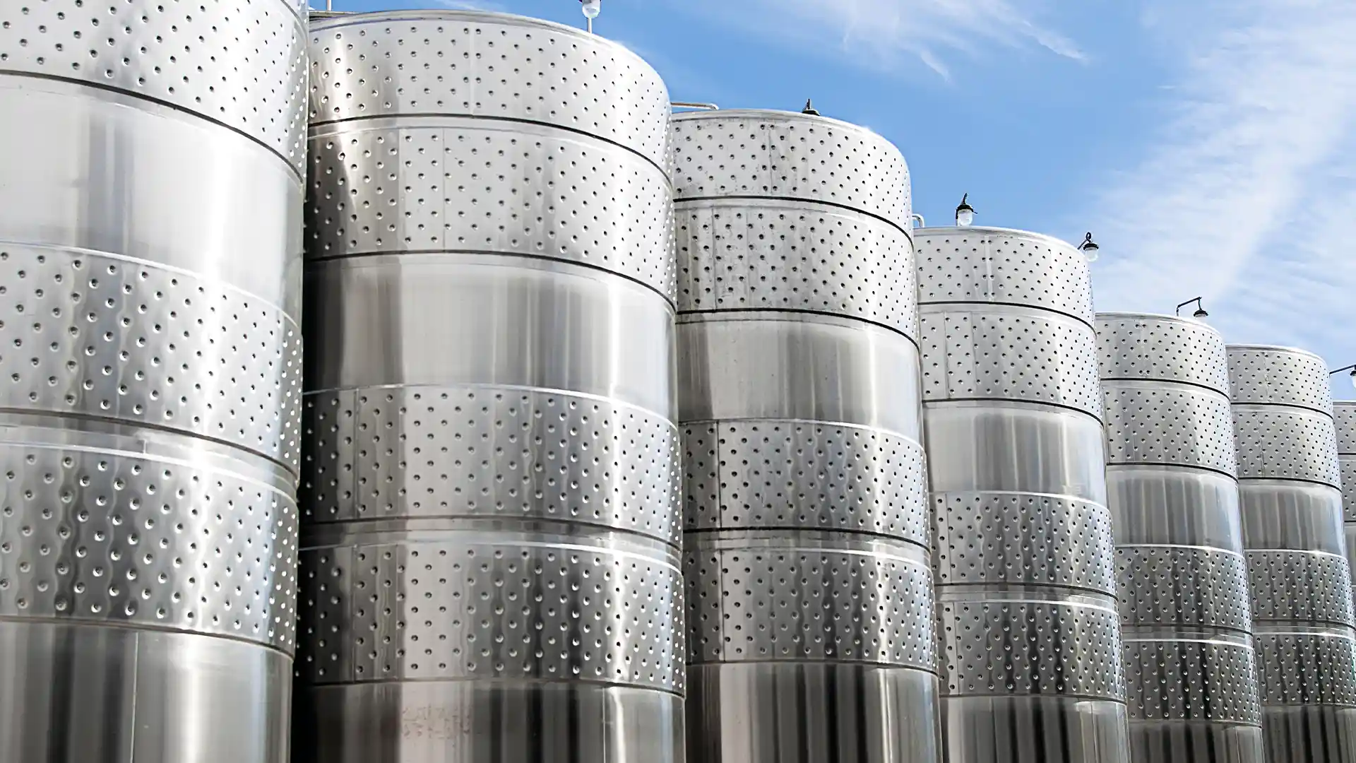 Row of large, shiny stainless steel fermentation tanks under a blue sky, conveying an industrial and modern tone, often found in wineries or breweries.
