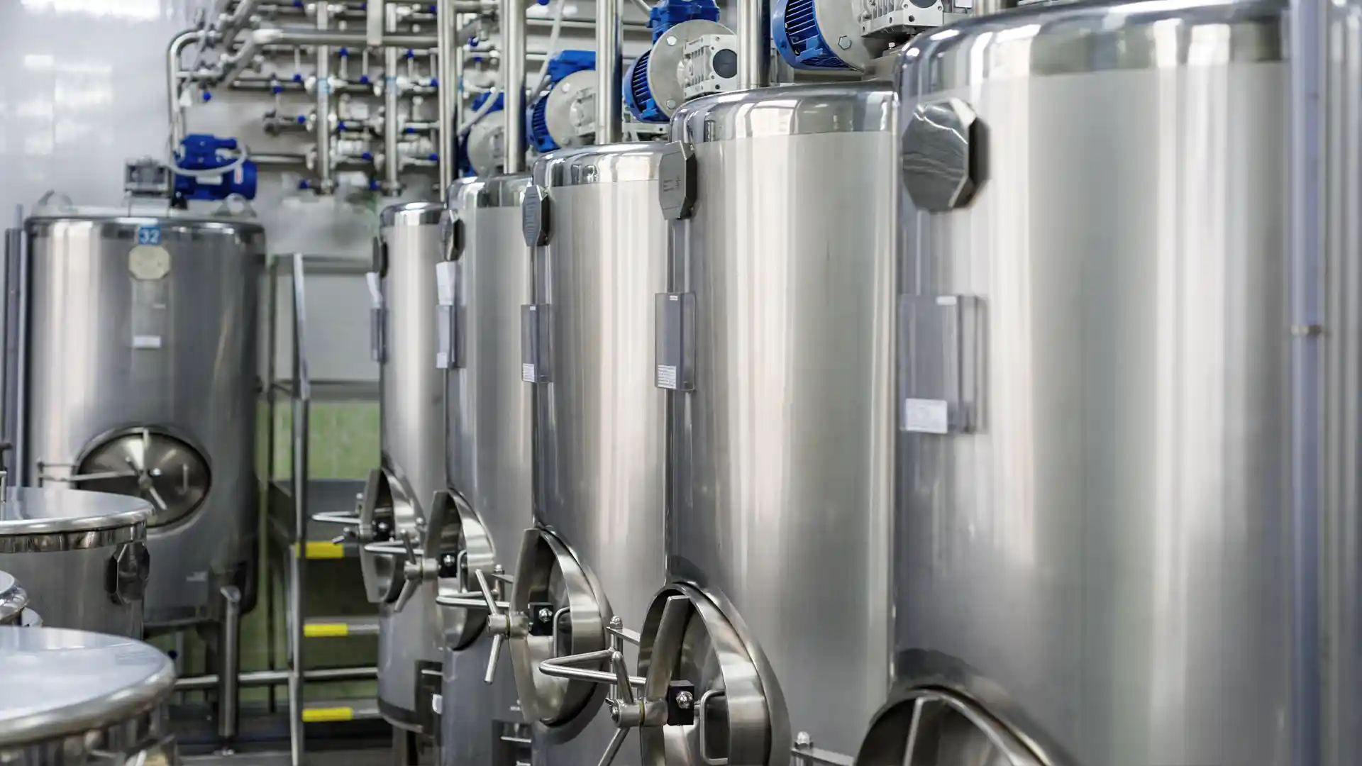 Row of shiny stainless steel fermentation tanks in a brewery, connected by pipes and valves, suggesting a clean and efficient industrial environment.