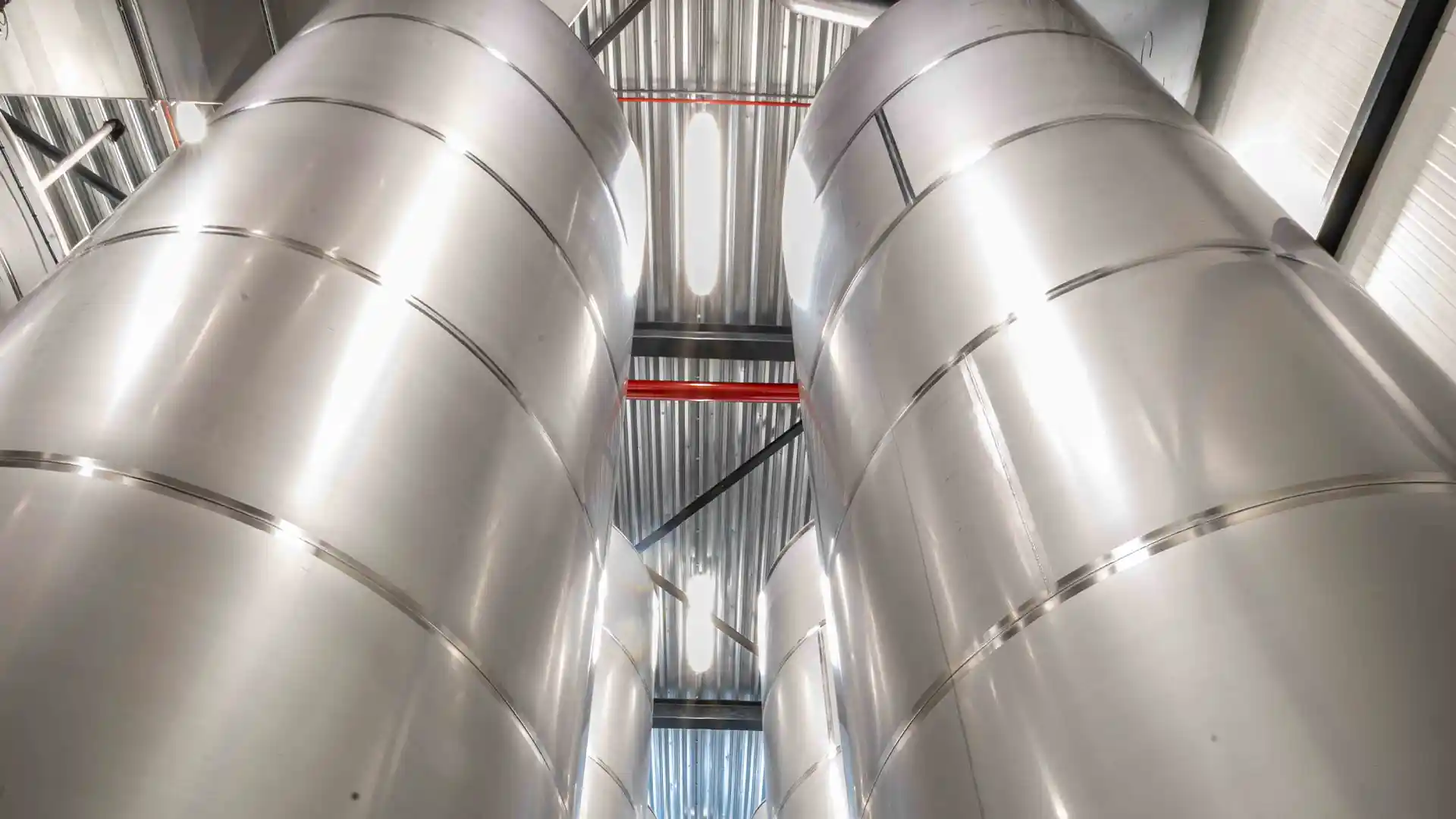 Four large stainless steel fermentation tanks in a brewery, towering upwards with a reflective surface. Overhead lights cast a bright, industrial look.