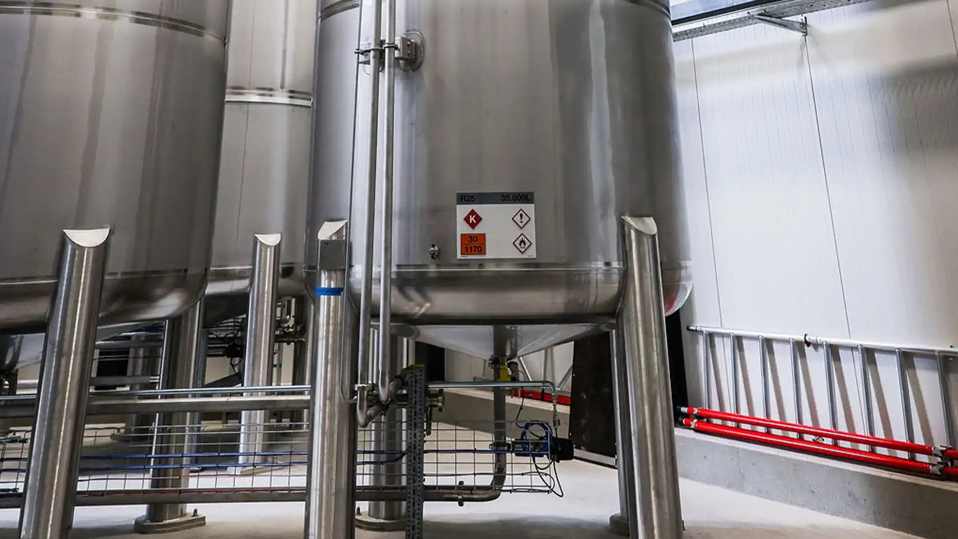 Large stainless steel industrial tanks with warning labels are in a bright, clean facility. Pipes and safety measures indicate a controlled environment.