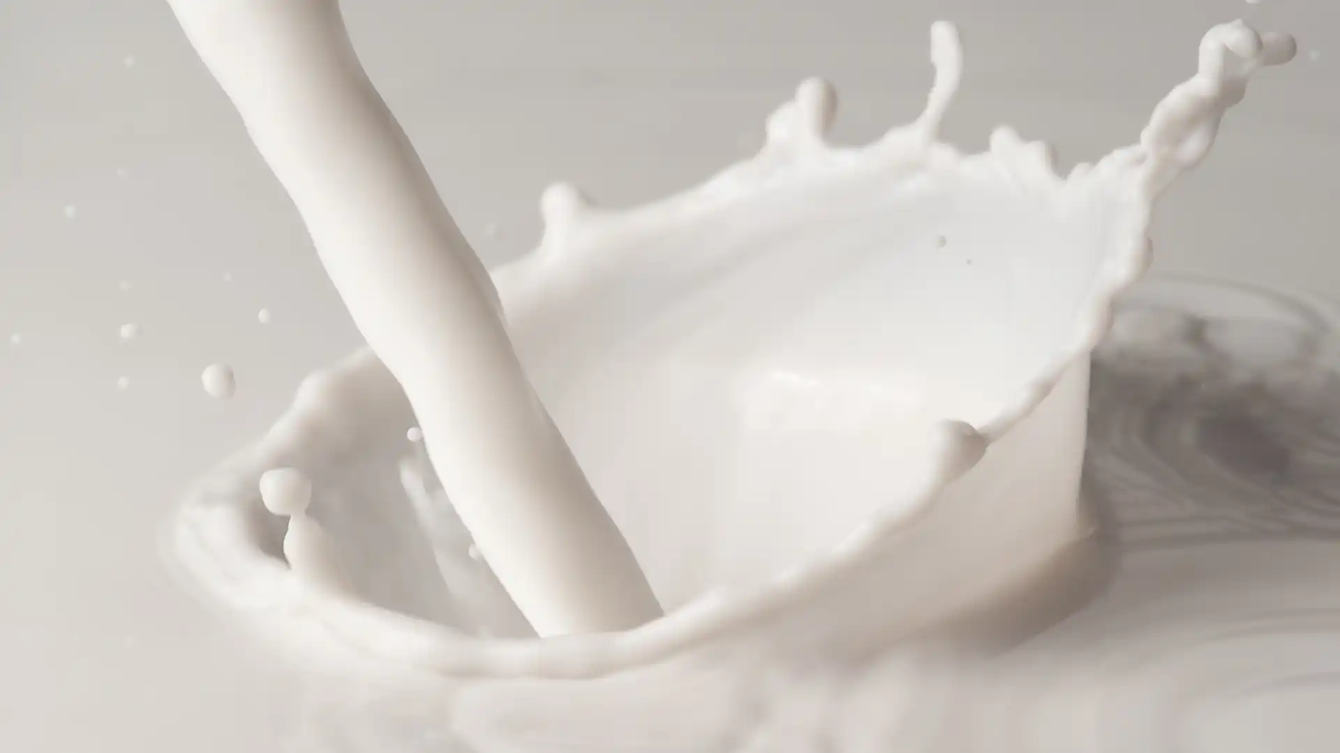 A stream of milk splashes into a creamy pool, creating a smooth, rippling wave and droplets. The image conveys freshness and fluidity.