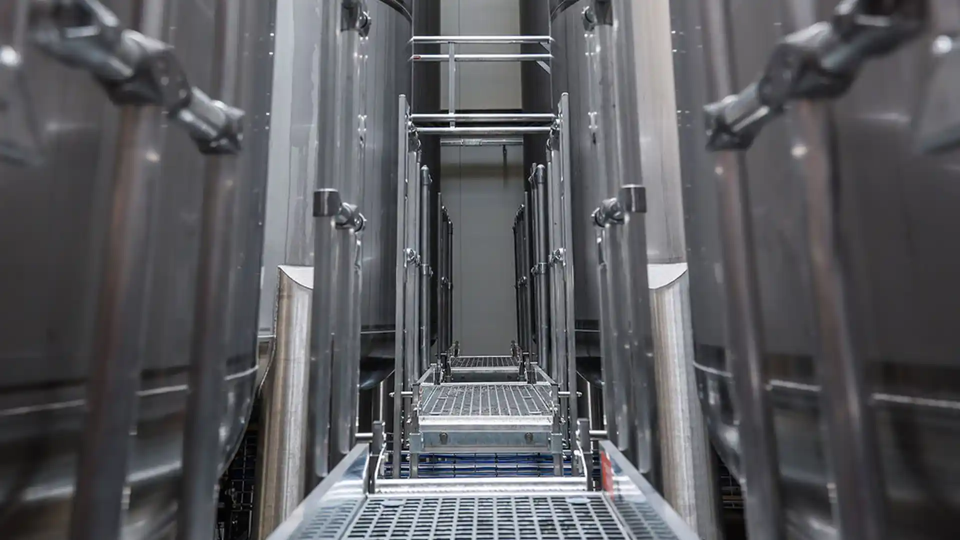 A narrow metal walkway flanked by large industrial tanks, conveying a sense of symmetry and industrial efficiency in a factory setting.