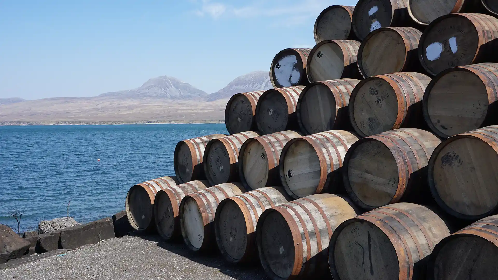 A stack of wooden barrels is arranged on a shore with a scenic backdrop of a calm blue sea and distant mountains under a clear sky.