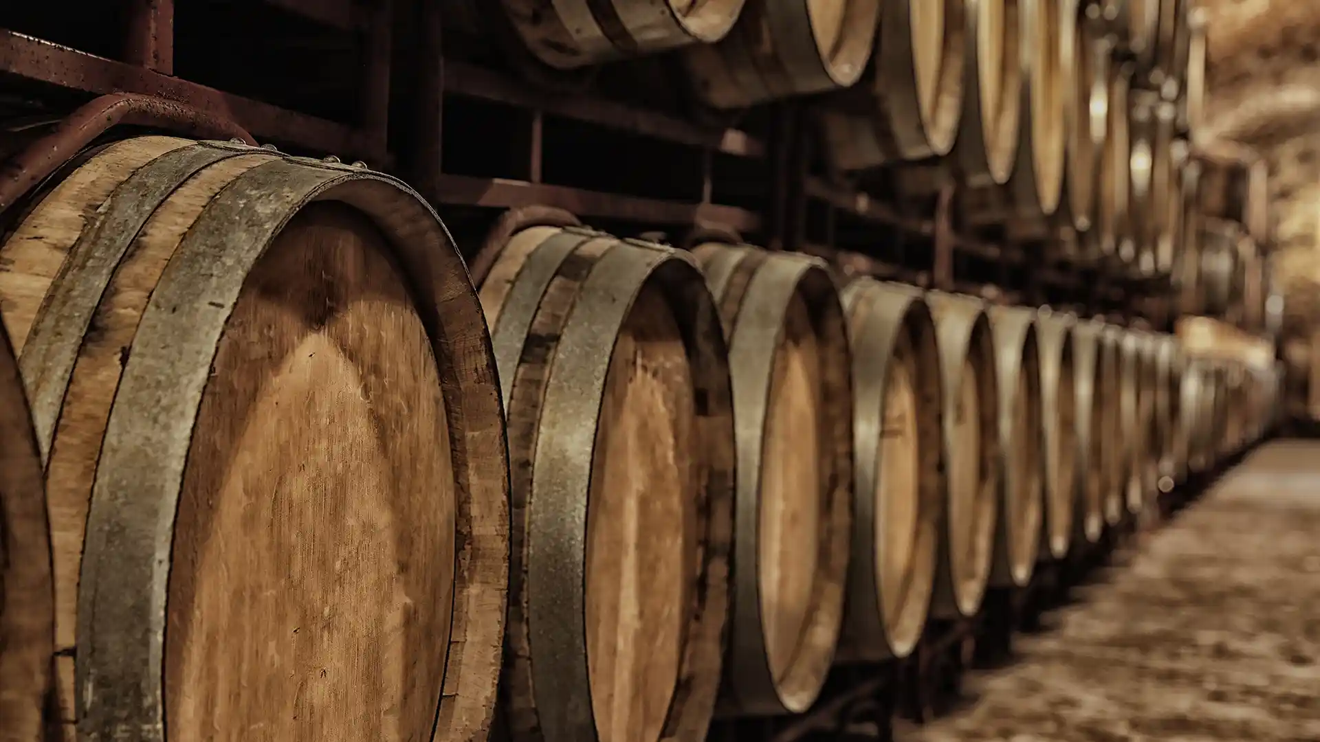 Rows of wooden wine barrels with metal hoops are stacked in a dimly lit cellar, creating an atmosphere of rustic elegance and tradition.