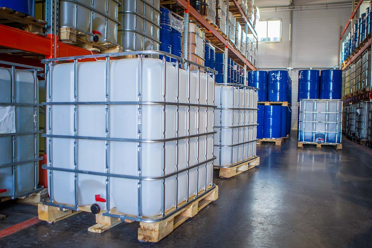 A warehouse interior with large white containers on pallets and blue barrels stacked on shelves. The scene is well-lit, clean, and organized.