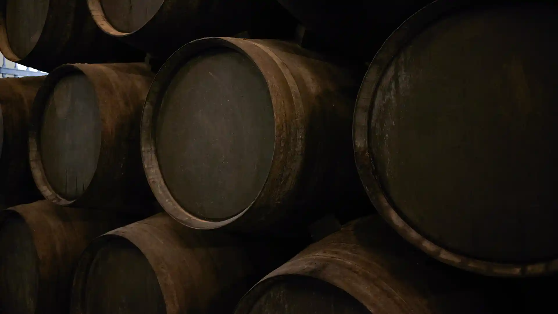 A stack of large, weathered wooden barrels in dim lighting, conveying an atmosphere of age and tradition, likely in a winery or distillery.