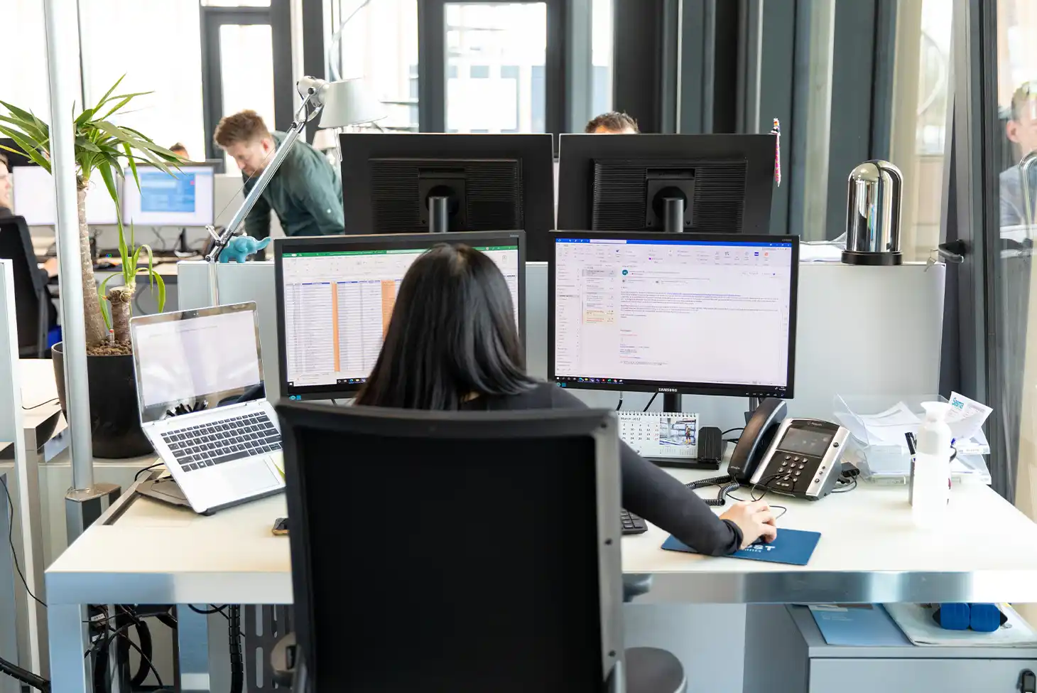 A woman with long hair works at a dual-monitor setup in a modern office. Nearby are a laptop, phone, and potted plant. The atmosphere is busy yet focused.