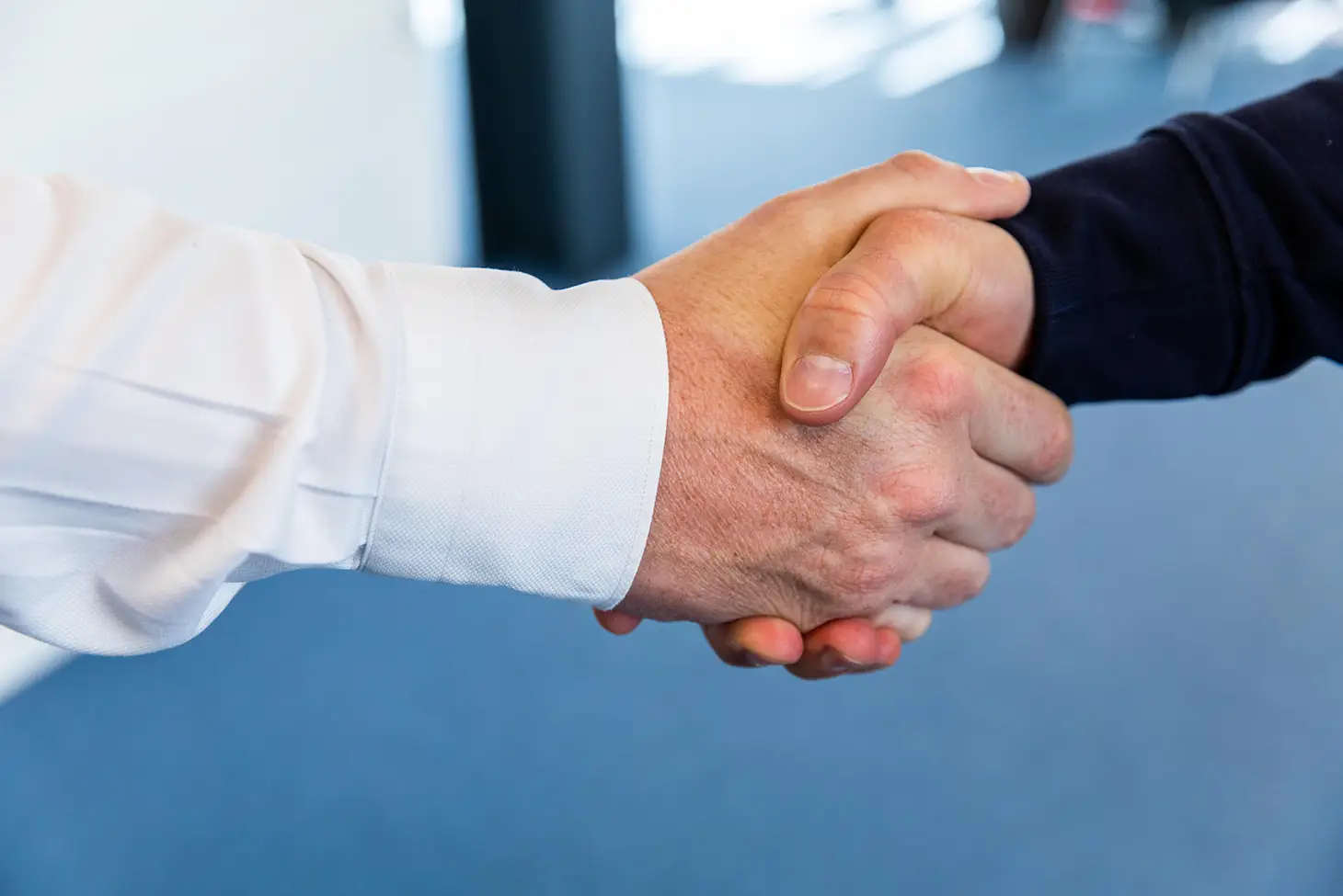 Two people are shaking hands in a professional setting, with one wearing a white shirt and the other in a dark sweater, suggesting agreement or partnership.