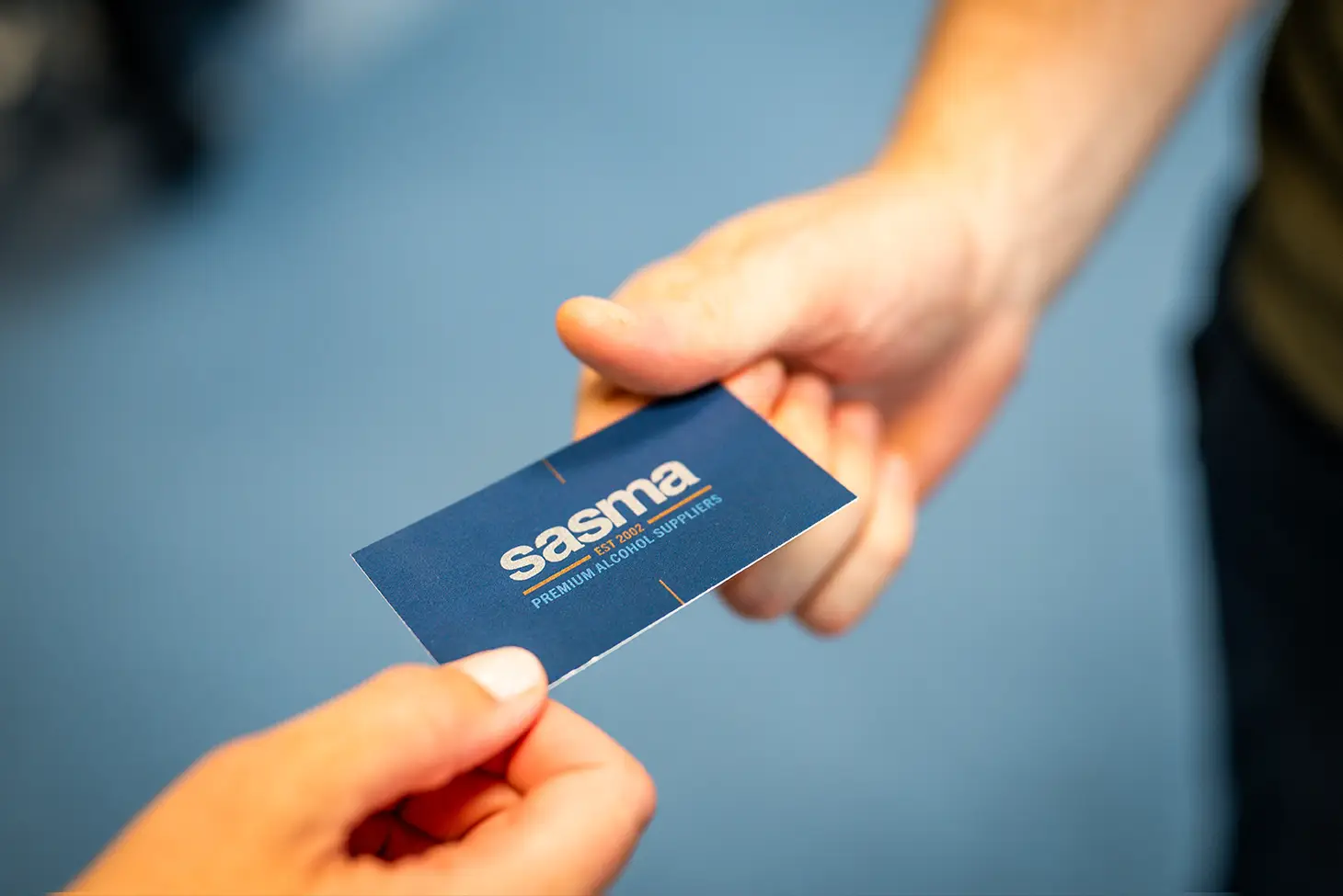 Two hands exchange a blue business card labeled "Sasma," conveying a professional interaction. The background is softly blurred in light blue.
