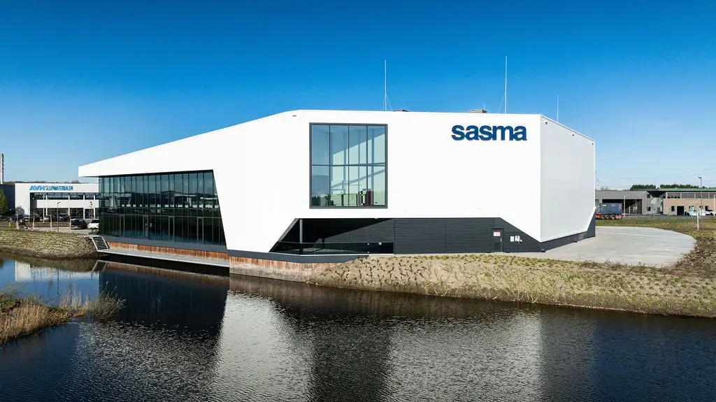 Modern white building with large windows, labeled "sasma," stands beside a calm body of water under a clear blue sky.