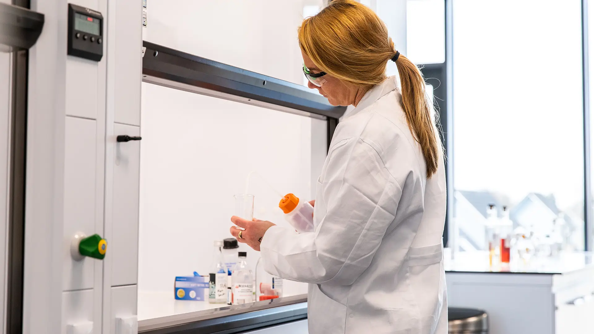 A woman in a lab coat works at a lab bench, holding a bottle. She's surrounded by various scientific equipment, conveying focus and precision.