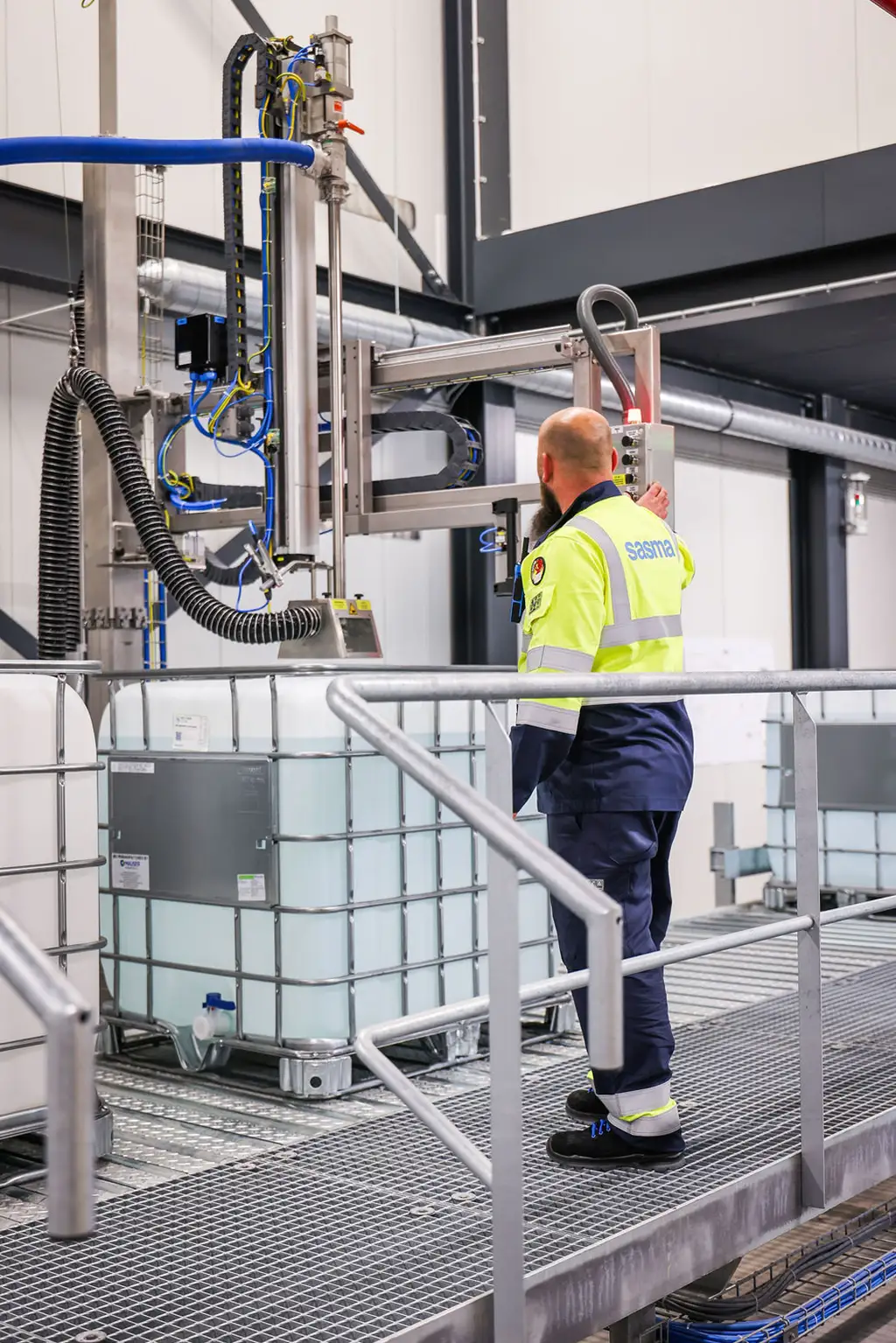 A worker in a high-visibility uniform operates industrial equipment in a factory. He stands near large liquid containers, adjusting a machine with hoses.
