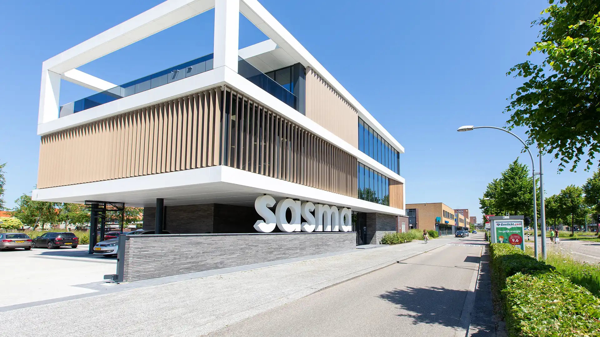 Modern three-story building with a white and beige facade, large windows, and the word "Sasma" displayed prominently. Clear blue sky backdrop.