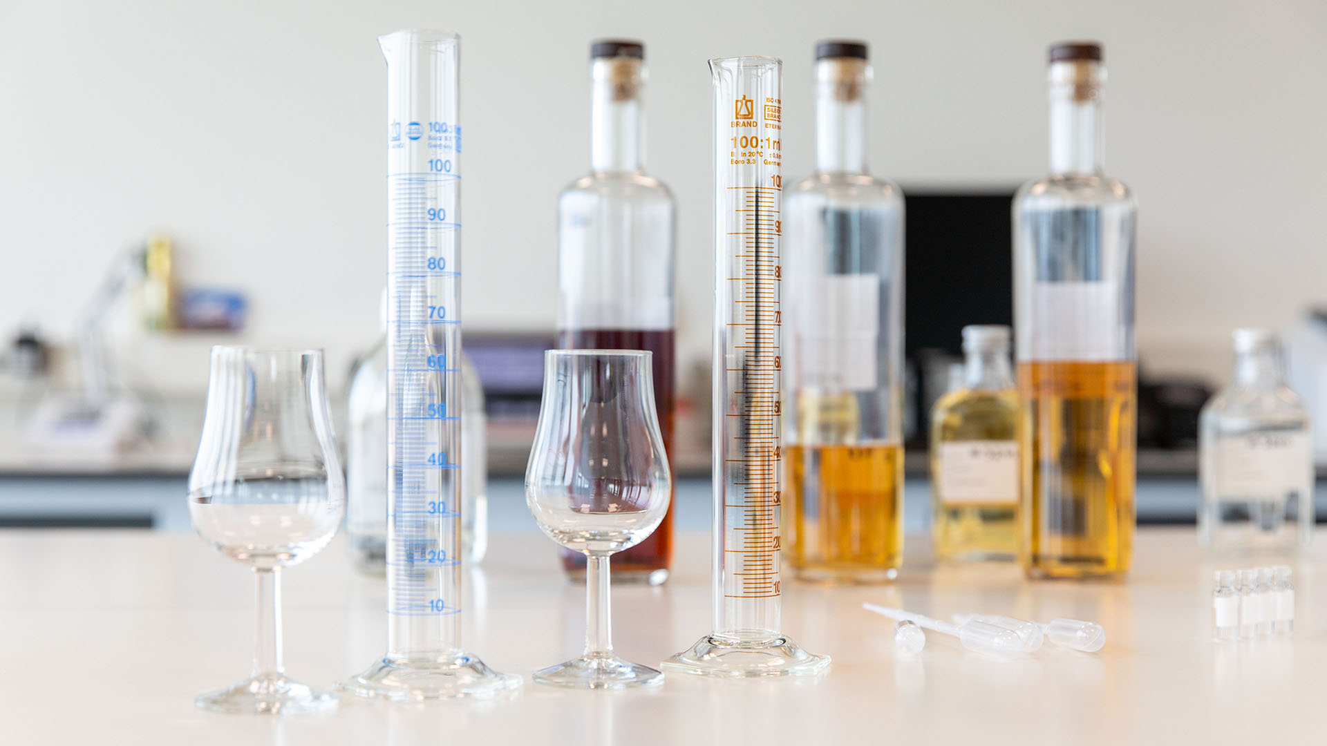A laboratory setting with graduated cylinders and a tasting glass in the foreground. Blurred liquor bottles with various shades in the background.