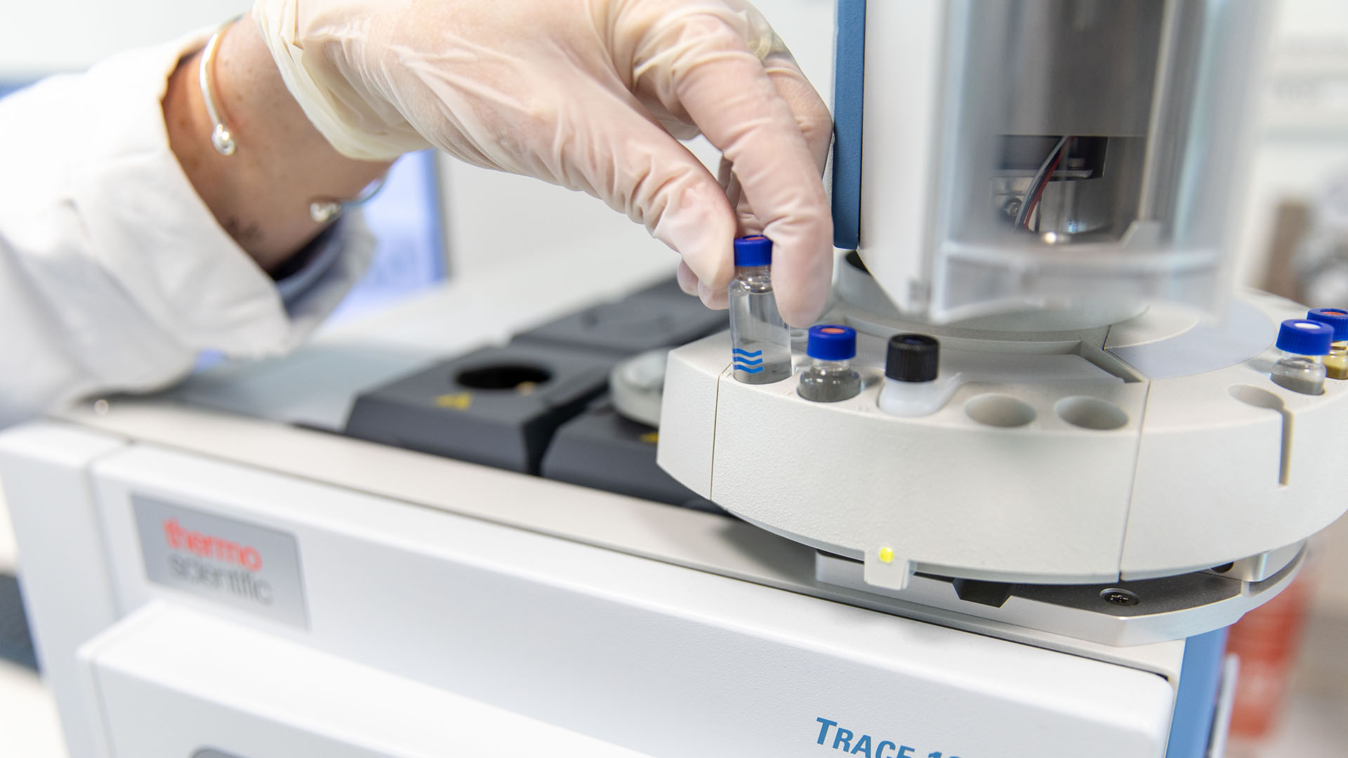 A gloved hand places a vial into a scientific analyzer labeled Trace 1300. The setting is a lab, conveying a sense of precision and advanced technology.