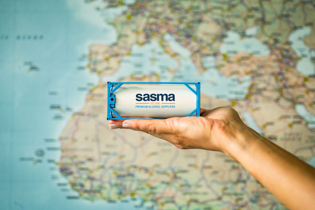 A hand holds a miniature shipping container with "sasma" branding in front of a blurred world map, indicating global supply or logistics.
