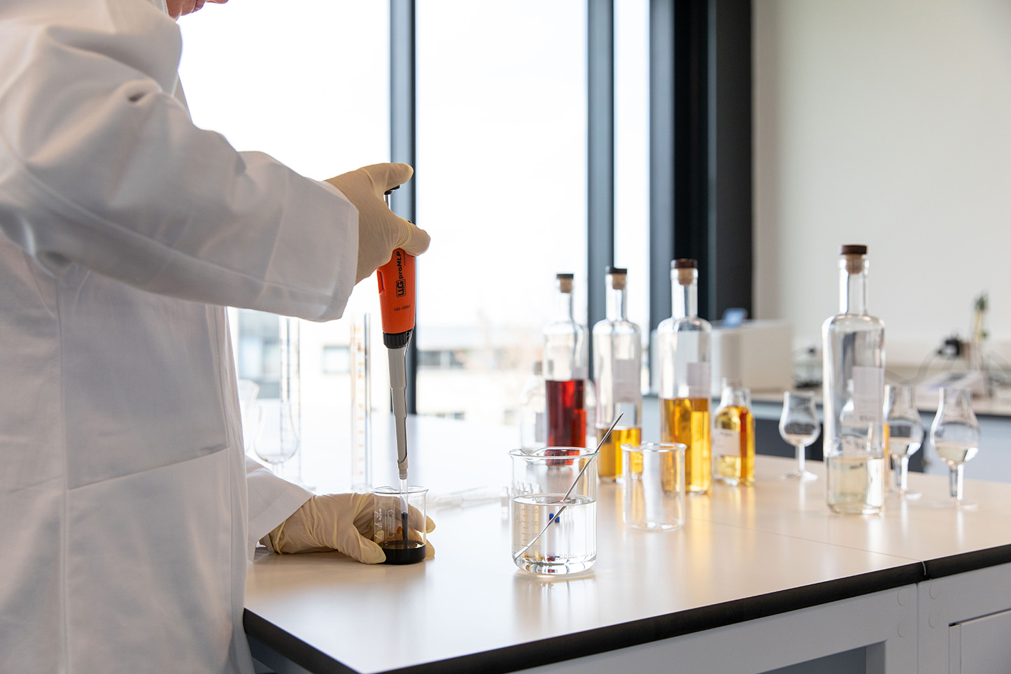 A person in a lab coat uses a pipette to transfer liquid into a glass beaker on a counter with bottles of various colored liquids against large windows.