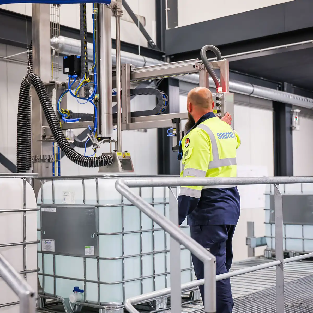 A worker in a high-visibility uniform operates industrial equipment in a factory. He stands near large liquid containers, adjusting a machine with hoses.