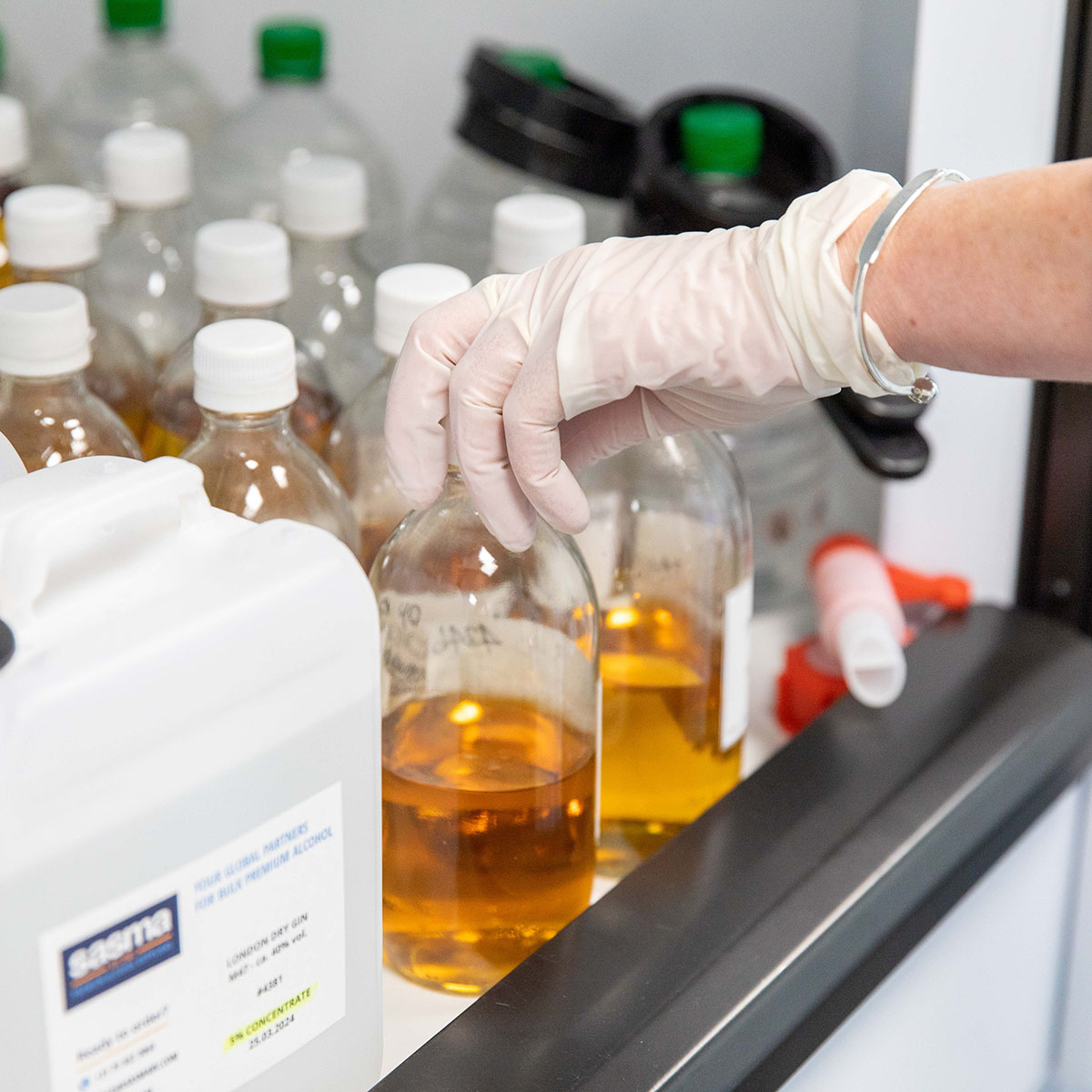 A gloved hand handles a glass bottle with amber liquid in a lab, surrounded by other bottles and containers. The scene is clean and sterile.