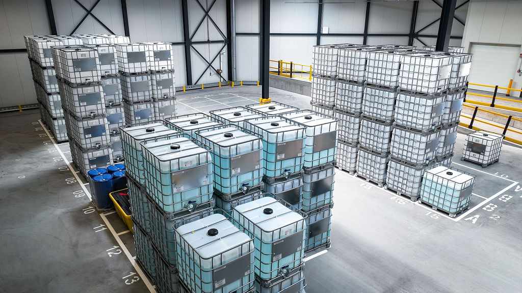 Stacks of white storage containers in a warehouse, organized neatly on a gray floor. Blue barrels are visible to the side, creating an industrial vibe.