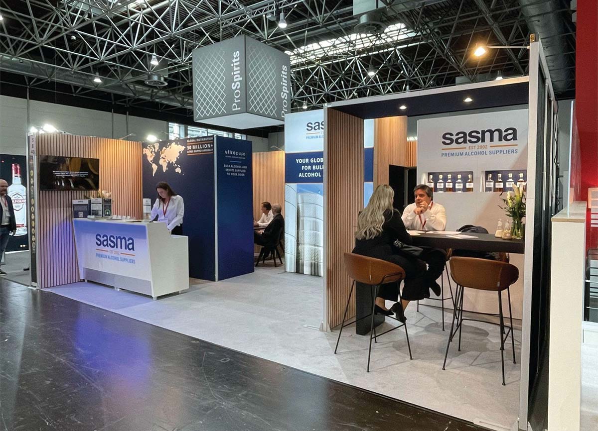 Trade show booth with "Sasma" branding, featuring a reception desk and seating area. People engage in discussions, creating a professional and welcoming atmosphere.