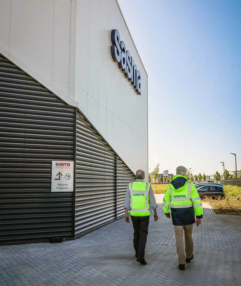 Two people in high-visibility vests walk toward a commercial building labeled "Sasma" under a clear blue sky.