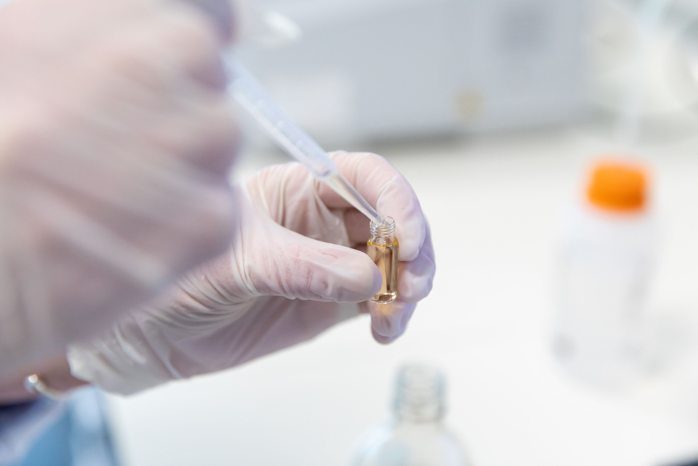 A gloved hand uses a dropper to transfer liquid into a small vial in a laboratory setting, conveying precision and scientific focus.