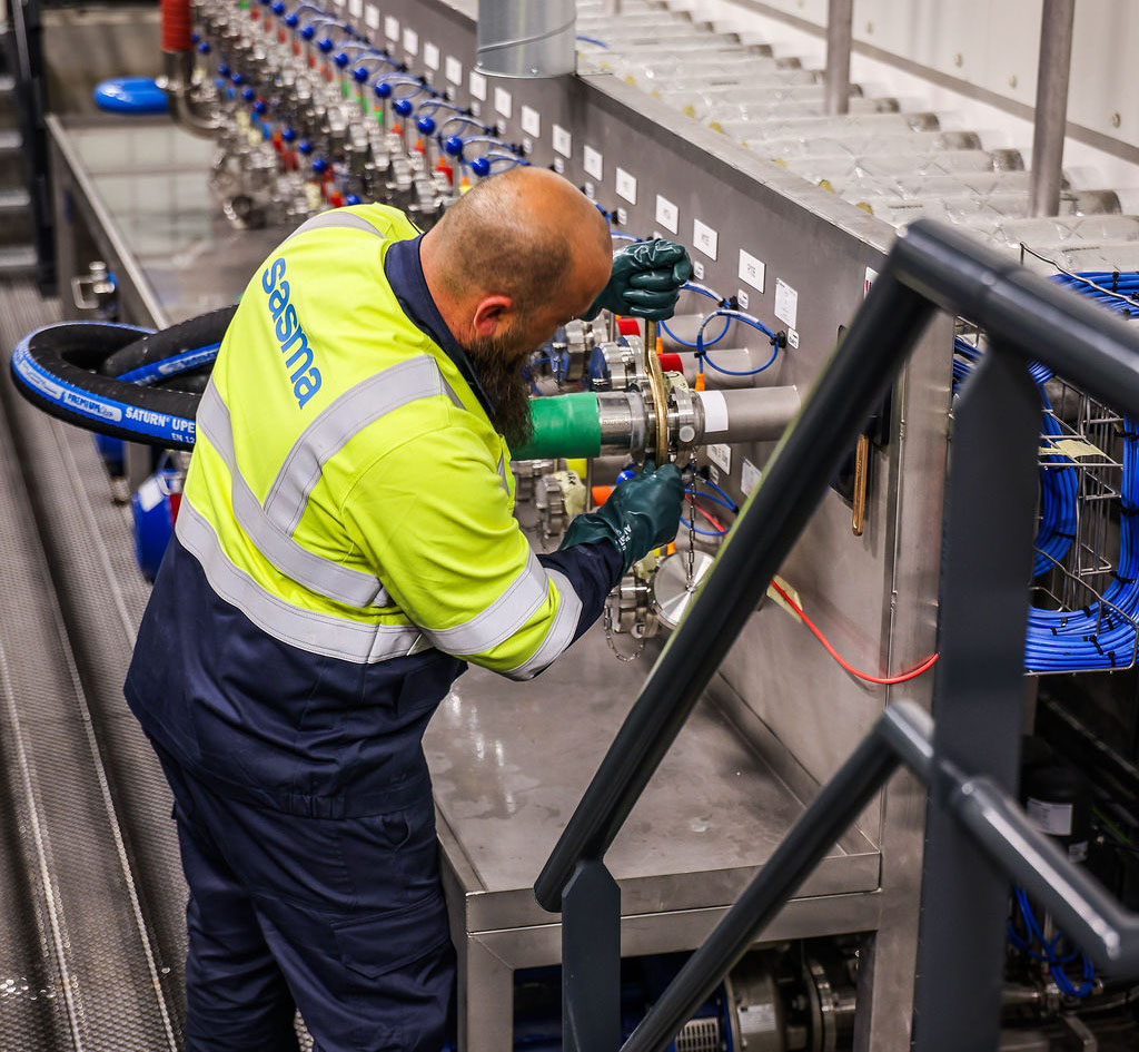 A worker in a high-visibility jacket adjusts machinery in an industrial facility. An array of pipes and colored cables is visible on a metal platform.