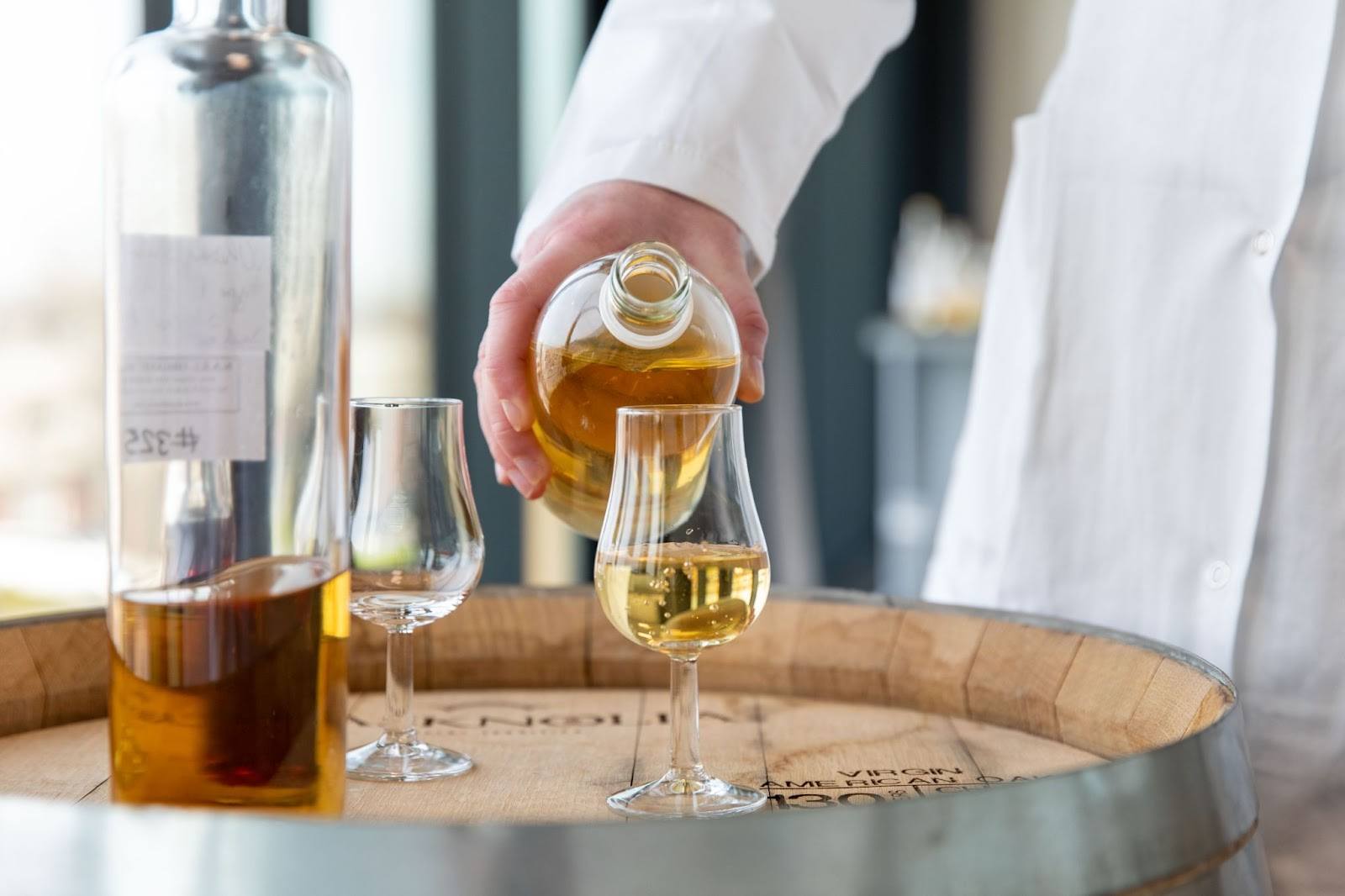 A person in a white coat pours whisky, into a glass on a wooden barrel. Nearby, a bottle and two more glasses are visible, suggesting tasting.